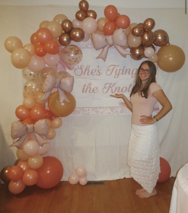 Home 10 A bride-to-be standing before a peach balloon garland and a She's Tying the Knot backdrop