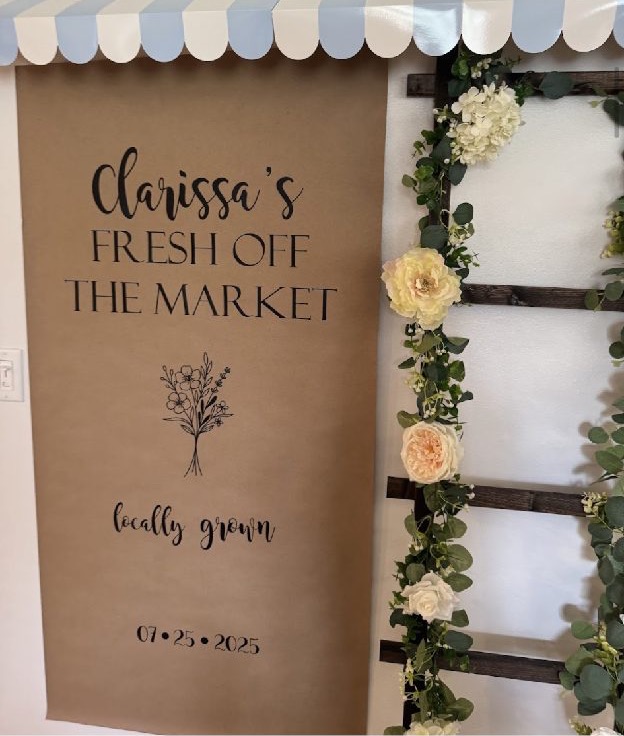 Home 11 A hand-lettered kraft market sign framed by white florals and greenery at a bridal shower